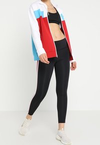 Felpa rossa, bianca e blu con zip abbinata a leggings neri con strisce rosa sui lati. Indossata con scarpe sportive grigio chiaro.