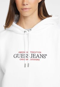 Biela mikina s kapucňou a šnúrkou, s vyšitým logom, na ktorom je nápis "GUESS JEANS" v čiernom a červenom texte. Mäkká textúra látky.