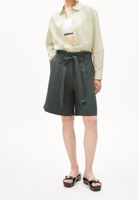 Světle zelená košile s dlouhým rukávem přes bílý crop top, doplněná tmavě zelenými šortkami s vysokým pasem, které mají uvázaný pásek, a plochými černými sandály.