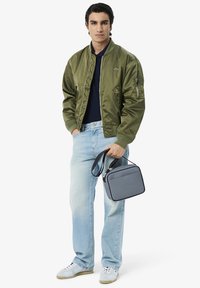 Giubbotto bomber verde oliva, polo blu navy, jeans chiari in denim, borsa a tracolla grigia con logo in rilievo, sneakers bianche. Outfit casual e moderno.