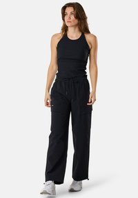 Zwarte ribbel tanktop gecombineerd met losse zwarte cargobroek met zakken. Het model draagt witte sportschoenen.