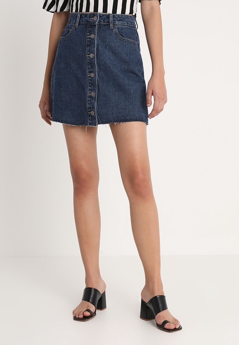 Falda mini de mezclilla, azul oscuro, con cierre de botón en la parte delantera, ligero deshilachado en el dobladillo, con dos bolsillos delanteros, combinada con sandalias de tacón negras.