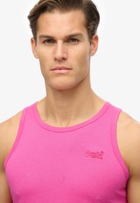 Top de algodón rosa sin mangas con un cuello redondo. Presenta un logo bordado en el pecho, textura suave y forma ajustada.