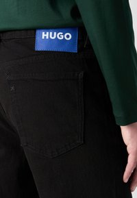 Gros plan d'une personne portant un jean noir avec une étiquette de marque HUGO bleue sur la ceinture et une chemise à manches longues vert foncé.