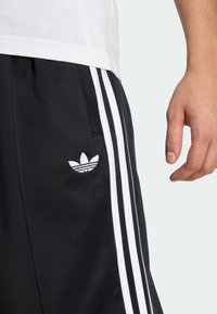 Crne sportske kratke hlače s bijelim prugama, s izvezenim bijelim logotipom. Tkanina je glatke teksture. Vidljiva je ruka koja drži tkaninu.