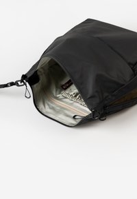 Schwarze Nylon-Tasche mit einem Reißverschluss-Öffnung. Das Innere weist eine helle Fütterung und eine kleine Tasche mit Reißverschluss auf. Details umfassen verstellbare Beschläge.
