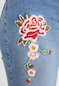 Vaqueros de mezclilla azul con intrincados bordados florales en rojo, crema y verde en el lateral, con una superficie texturizada.