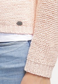 Pull en tricot rose clair avec un motif texturé, ourlet côtelé et une petite étiquette métallique de marque près du bas. Porté par-dessus un haut blanc.