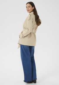 Beige trench coat s páskem v pase, knoflíkovými detaily a ramenními epauletami, v kombinaci s volnými modrými džínami a černými botami na podpatku.
