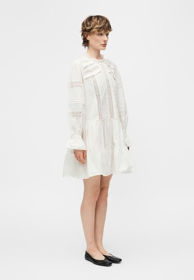 Γυναίκα που φοράει λευκό φόρεμα με μακριά μανίκια και δαντελωτές λεπτομέρειες, με γιακά και μανσέτες με βολάν, συνδυασμένο με μαύρα επίπεδα παπούτσια, στεκόμενη πλάγια.