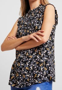 Svart blommig blus med volangärmar, prydd med rosa och gula blommor, med draperad halsringning och lätt material.