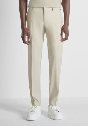 Beige op maat gemaakte broek gedragen met witte sneakers, getoond van taille tot voeten tegen een effen achtergrond.