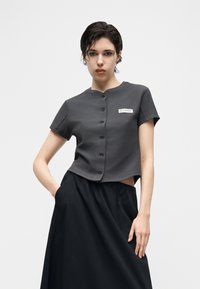 Νεαρή γυναίκα με κοντά σκούρα μαλλιά φοράει πουκάμισο σκούρου γκρι χρώματος με κοντά μανίκια και κουμπιά και μαύρη φούστα, ποζάροντας με το ένα χέρι στην τσέπη.