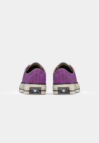Lilla lerrets-sneakers med lav profil, gummikappe foran og slitt hvit såle. Har svart og hvit merkeetikett på hælen.