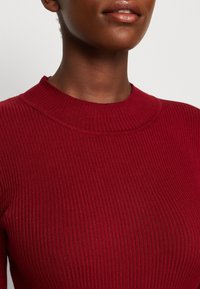 Gros plan sur une personne portant un pull côtelé à manches longues de couleur rouge foncé avec un col rond, montrant le bas du visage et le cou.