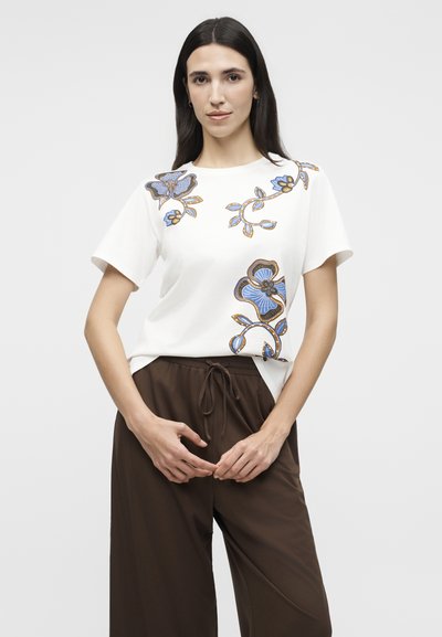 Γυναίκα που φοράει λευκό κοντομάνικο τοπ με γαλάζια και καφέ λουλουδένια σχέδια, σε συνδυασμό με χαλαρά καφέ παντελόνια με κορδόνι, στέκεται μπροστά σε απλό φόντο.