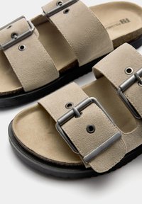 Beige Wildleder-Sandalen mit zwei breiten Riemen, metallischen Schnallen und einem strukturierten Fußbett. Schwarze Gummisohle mit einer leichten Kurve.