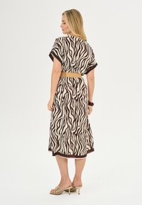 Maxi jurk met bruin en crèmekleurig zebra print. Korte mouwen, losse pasvorm en een strakke taille met een gouden riem. Draagt beige hoge hakken.