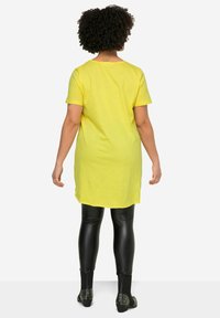 Camicia tunica gialla con maniche corte, design fluido, indossata sopra leggings neri lucidi, abbinata a scarpe nere con borchie argentate.