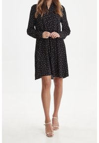 Abito nero con pois bianchi, maniche lunghe, chiusura frontale con bottoni e vita arricciata. Completato con una gonna a campana. Indossato con sandali con cinturini.
