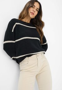 Sort oversized sweater med hvide vandrette striber, lavet af blødt materiale. Kombineret med lys beige tætsiddende bukser. Simpelt design, afslappet look.