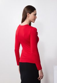 Červený top s dlhým rukávom vyrobený z elastického materiálu, s okrúhlym výstrihom a priliehavým strihom, skombinovaný s čiernou sukňou.
