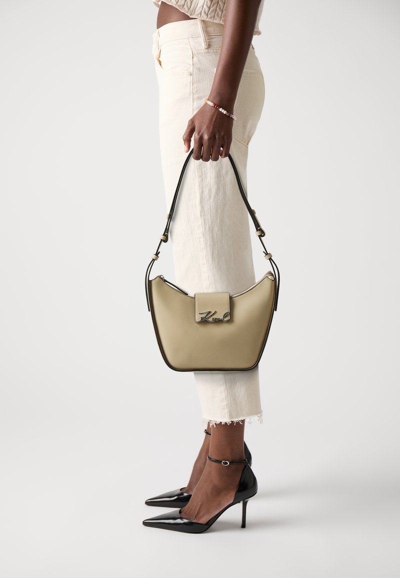 Beige håndveske med en buet design, sølvdetaljer og en logo. Modellen har på seg kremfargede bukser og svarte stiletthæl.