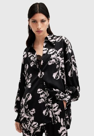 Chemise en satin noir avec un grand motif floral blanc, à manches longues, coupe oversized et fermeture par boutons. Présente des détails en dentelle en dessous.