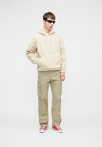 Sudadera de forro polar beige con bolsillo canguro y cordón. Combinada con pantalones cargo beige claro que tienen varios bolsillos. Zapatillas rojas.