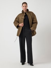 Femeie îmbrăcată cu o jachetă oversized maro, un pulover negru cu textură, pantaloni negri cu picioare largi și tocuri negre ascuțite, stând în fața unui fundal alb.