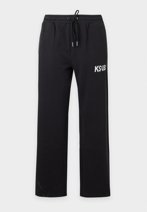 Jeune homme portant des lunettes de soleil noires, un gilet noir sur un T-shirt blanc, un pantalon noir avec un logo blanc et des bottes marron à lacets rouges, debout devant un fond blanc.