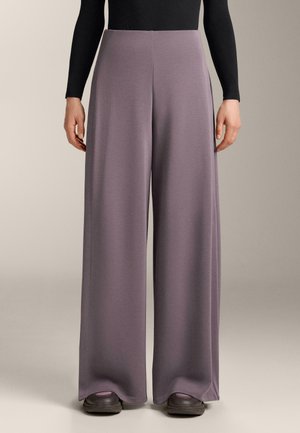 Osoba u visokim širokim hlačama u boji mahagonija kombiniranim s crnom dugom majicom i tamnim tenisicama, stoji ispred jednostavne pozadine.