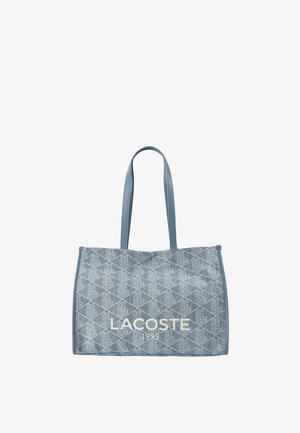 Blauwe denim tote bag met ruitpatroon, twee schouderbanden, knoopsluiting en "LACOSTE 1933" gedrukt op de voorkant.