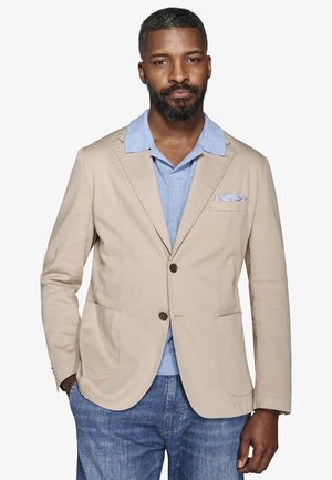 Homme barbu portant un blazer beige sur une chemise à col bleu clair et un jean bleu, debout avec une main dans la poche.