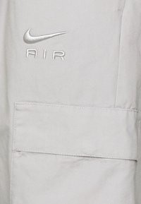 Světlá šedá tkanina se vyšitým logem Nike Air nad velkým obdélníkovým klopou kapsy.