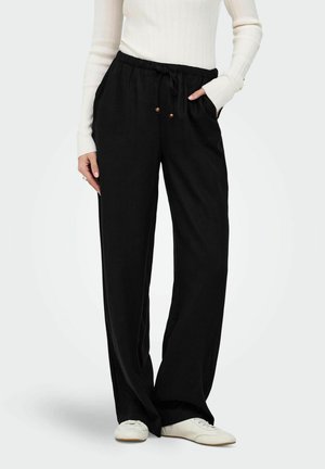 Nő, laza, fekete zsinóros nadrágot visel zsebekkel, fehér bordázott hosszú ujjú felsőt és fehér alkalmi cipőt egyszerű háttér előtt.