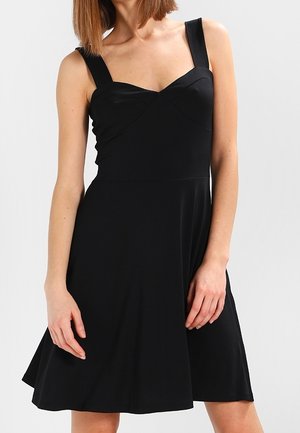 Kobieta w czarnej, bezrękawnikowej sukience z szerokimi ramiączkami i dopasowanym gorsetem, stojąca na tle jasnego, jednolitego tła.