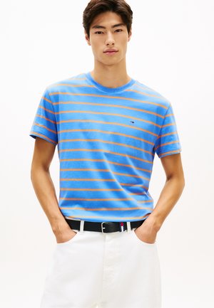 Νεαρός άνδρας που φοράει μπλε και πορτοκαλί ριγέ μπλουζάκι με μικρό λογότυπο, άσπρο παντελόνι, μαύρη ζώνη, χέρια στις τσέπες, ουδέτερο φόντο.