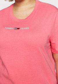 Camiseta de algodón rosa con cuello redondo, con texto bordado en blanco "TOMMY JEANS" y una pequeña franja de logo en rojo, blanco y azul.