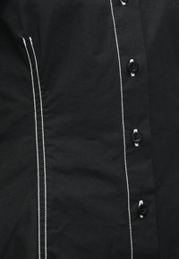 Tissu noir avec une double couture verticale blanche à gauche et des boutons noirs cousus avec du fil blanc sur le côté droit.