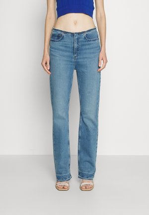 Jean slim fit en denim bleu porté par une personne avec un top bleu court et des sandales blanches à brides, debout devant un fond uni.