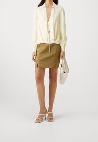 Blusa crema con maniche a sbuffo, mini gonna verde oliva con spacco laterale, sandali bianchi con tacco alto e una piccola borsa bianca.