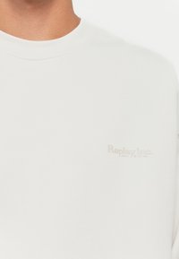 Sudadera blanca con cuello redondo, hecha de tela suave, que presenta un sutil logo gris "Replay Inc." en el pecho.