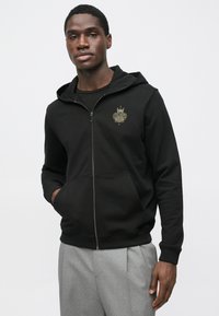 Camisola preta com fecho zip feito com tecido de mistura de algodão. Apresenta bolsos frontais, punhos canelados e um detalhe de logótipo dourado no peito.