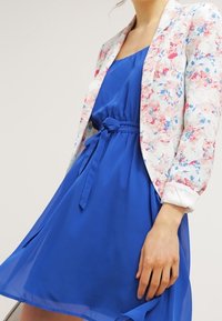 Femme portant une robe bleu royal avec une ceinture nouée à la taille et un blazer floral blanc avec des fleurs roses et bleues, manches retroussées.