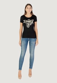 T-shirt nera a maniche corte con una grafica triangolare che presenta foglie di palma e dettagli metallici, abbinata a jeans skinny azzurri chiari e tacchi nudi.