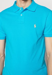 Tyrkysová polo tričko vyrobené z texturované bavlny, s límečkem, dvěma knoflíky a malým vyšitým logem na hrudi.