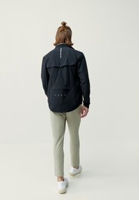 Svart lättviktsjacka med hög krage, dragkedjestängning och reflexdetaljer; kombinerat med ljusgröna åtsittande byxor och vita sneakers.