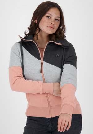 Jonge vrouw met sproeten draagt een kleurblok hoodie met ritssluiting in zwart, grijs en perzik, staande tegen een effen witte achtergrond.