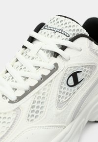 Λευκό αθλητικό παπούτσι με πλέγμα σε πάνελ, υφές από συνθετικό υλικό και γκρι λεπτομέρειες. Διαθέτει λευκά κορδόνια και μαύρο λογότυπο στο πλάι.